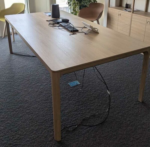 Table de réunion en bois couleur chêne clair, plateau rectangulaire à bords arrondis avec 2 trappes passe-câbles. 76 x 240 x 120 cm. Localisation : 6 ouest