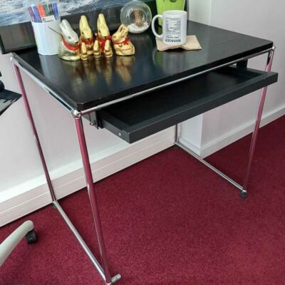 Table de marque USM HALLER, plateau en bois de couleur noire avec une tablette rétractable en ceinture, reposant sur une structure en acier chromé. 70 x 76 x 54 cm. Localisation : Canada 1er (2B01)
