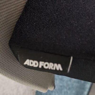 Fauteuil de bureau sur roulettes de marque Add’form, modèle Aura, assise en tissu noir et dossier en tissu gris clair, piètement en métal noir. Quantité disponible: 800 unités.
