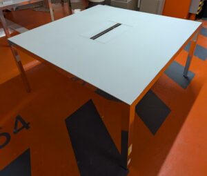 Table de réunion de forme carrée de marque Unifor, plateau en bois stratifié et verre avec trappe technique, piètement chromé. 74 x 150 x 150 cm. Localisation : 5 avenue Kleber - 75116 Paris